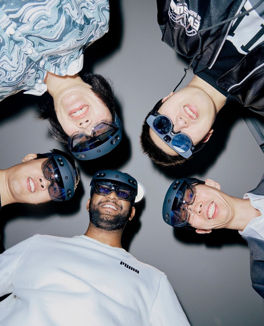 Five diverse adults gather indoors, wearing VR headsets and casual clothing, captured in a low-angle shot against a neutral gray background.