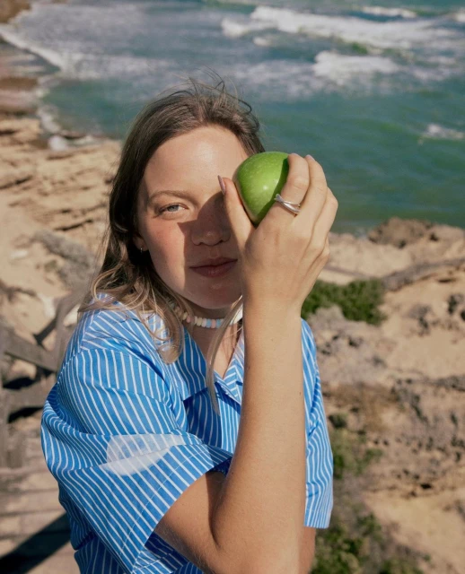 Young adult female poses with a green apple covering one eye outdoors near the ocean wearing a blue-and-white striped shirt.