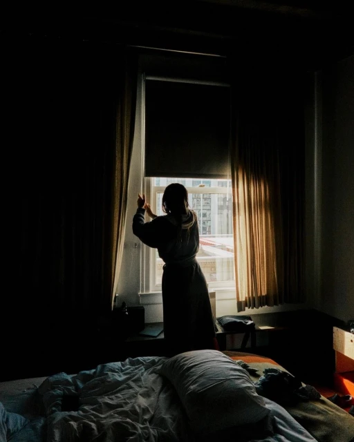 Woman opens curtains in a hotel room, revealing a city skyline in the background, with a bed in the foreground, during daytime.