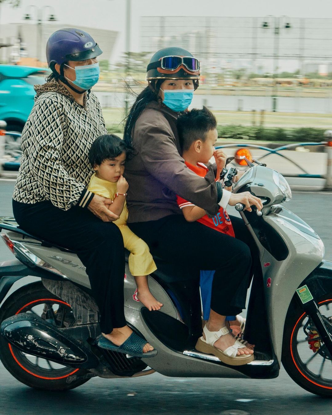 Four people, two adults and two children, ride together on a scooter in an urban area. All are wearing face masks.