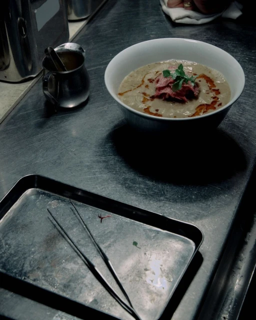 Overhead shot of a bowl of soup topped with meat and herbs, a metal tray with tweezers, and a metal pitcher on a metal surface.