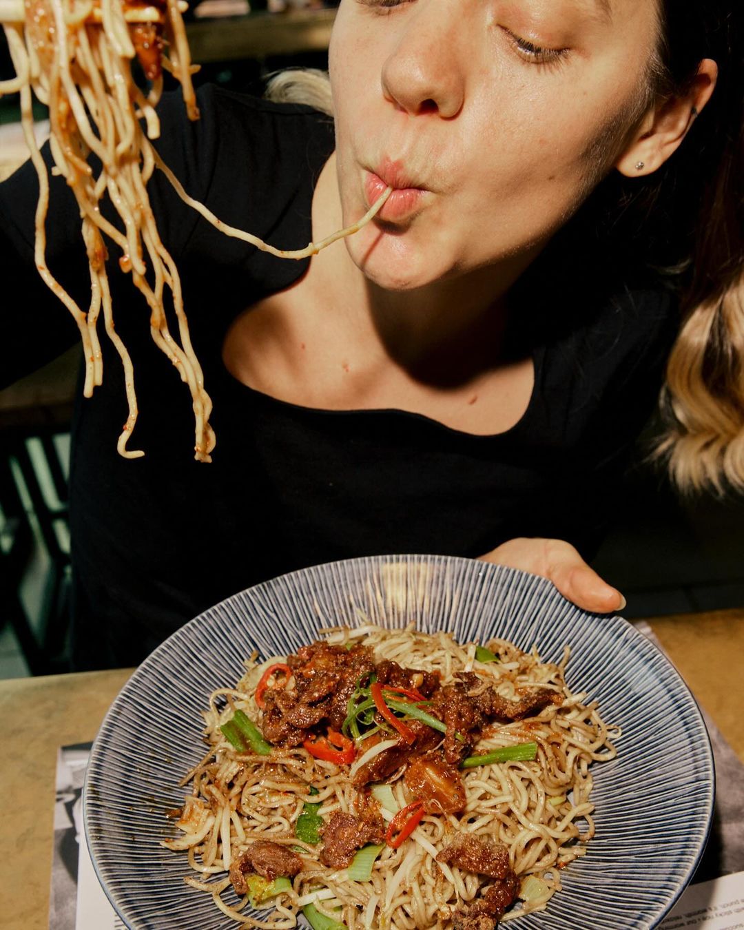 Blonde woman eats noodles with chop sticks over a bowl of noodles and meat with a dark background, taken in a low angle.