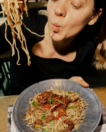 Blonde woman eats noodles with chop sticks over a bowl of noodles and meat with a dark background, taken in a low angle.