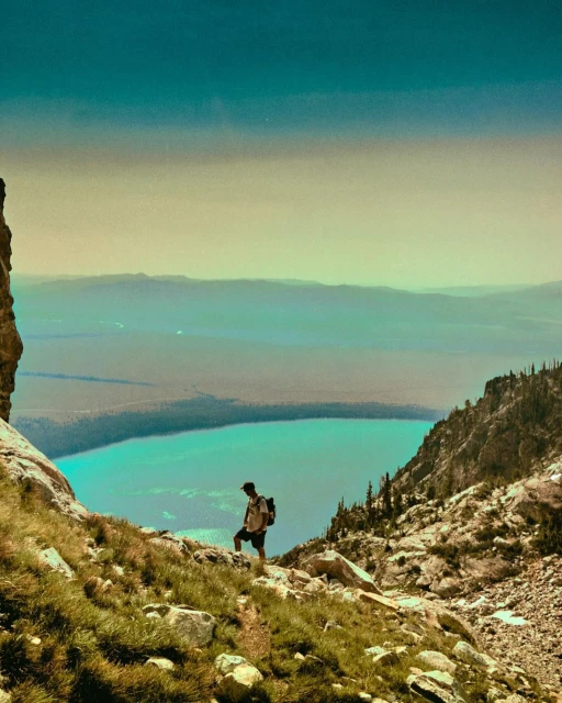 Adult male hiker with backpack overlooking turquoise lake and mountain range from a rocky, grassy slope on a clear sunny day.