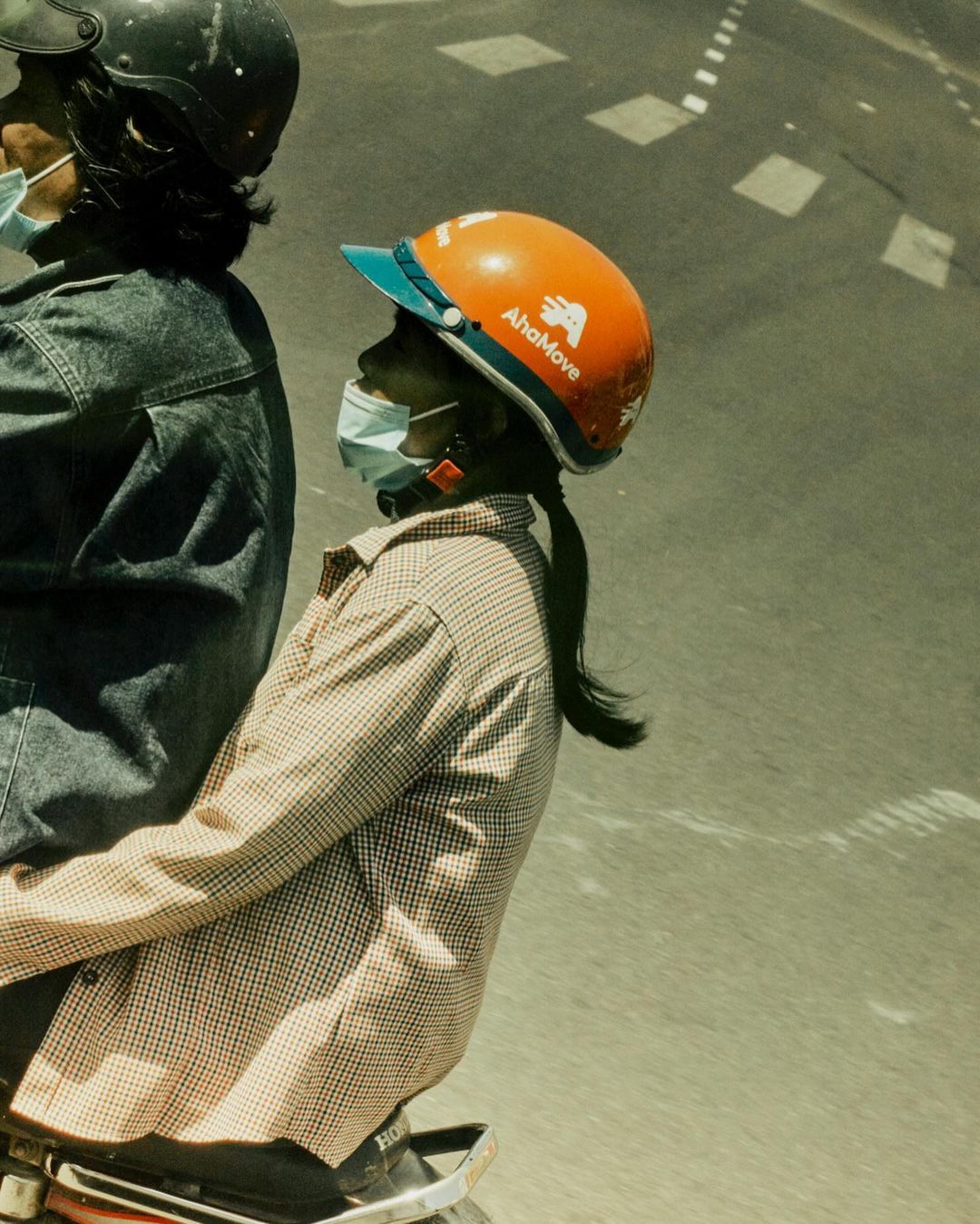 Two people wearing helmets and face masks ride on a motorcycle on a paved street. One person wears an orange safety helmet.