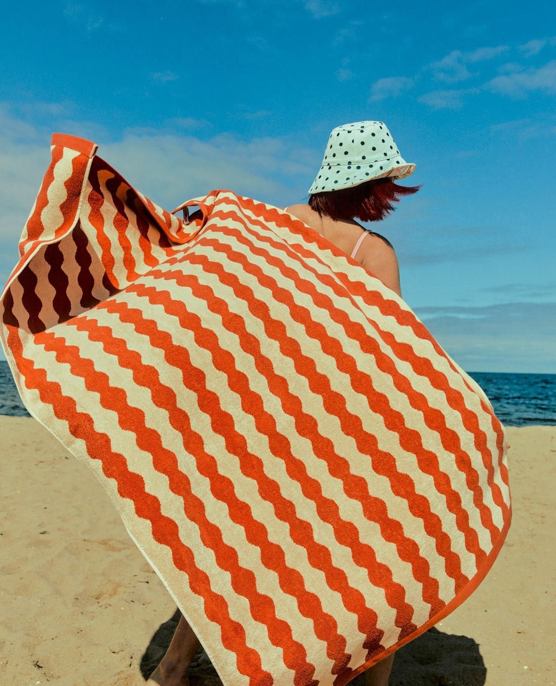 A woman wearing a polka dot bucket hat holds up a large orange and white patterned beach towel against the ocean and sandy beach background.