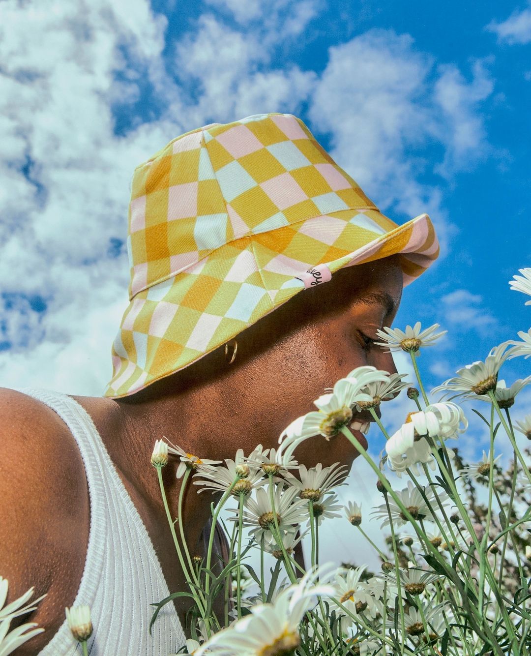 Person smelling daisies in a field, wearing a yellow and pink checkered bucket hat with a bright blue sky and clouds in the background.