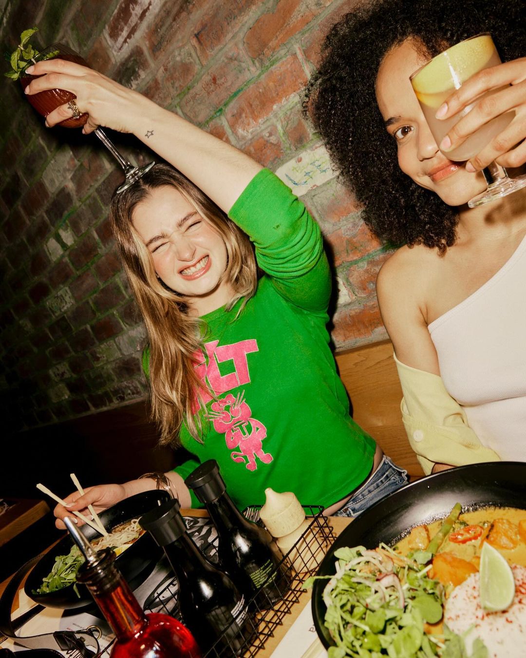 Two young women clink glasses with cocktails at a restaurant table with Asian food and drinks, illuminated by warm lighting and a brick wall backdrop.