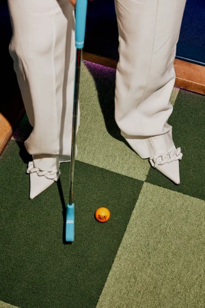 Woman in white pants and chain-trimmed shoes on a checkered green mini golf course, holding a blue golf putter near an orange golf ball.