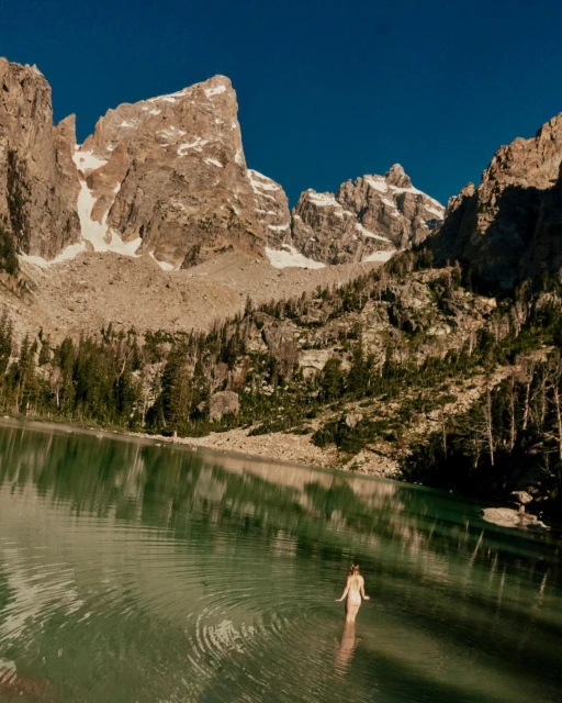 Woman wades into a tranquil lake with ripples, framed by majestic mountains and clear blue sky, creating a serene natural scene.