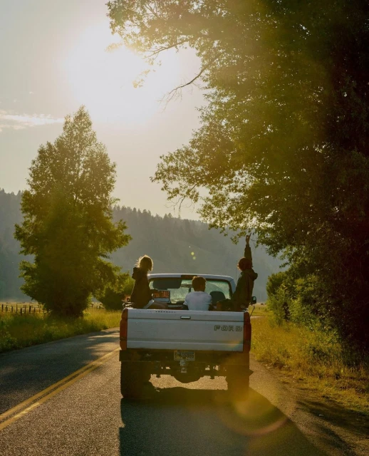 People ride in the back of a pickup truck on a rural road under a sunny sky, surrounded by trees and a mountain backdrop.