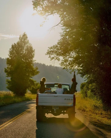 People ride in the back of a pickup truck on a rural road under a sunny sky, surrounded by trees and a mountain backdrop.