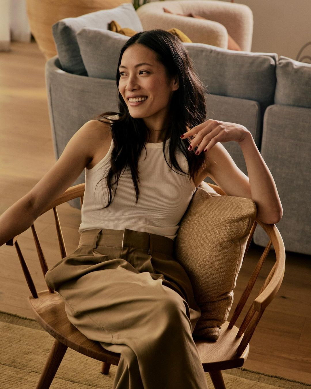 Indoor shot of an Asian woman in a white top and brown pants sitting in a wood chair and smiling with a couch in the background.