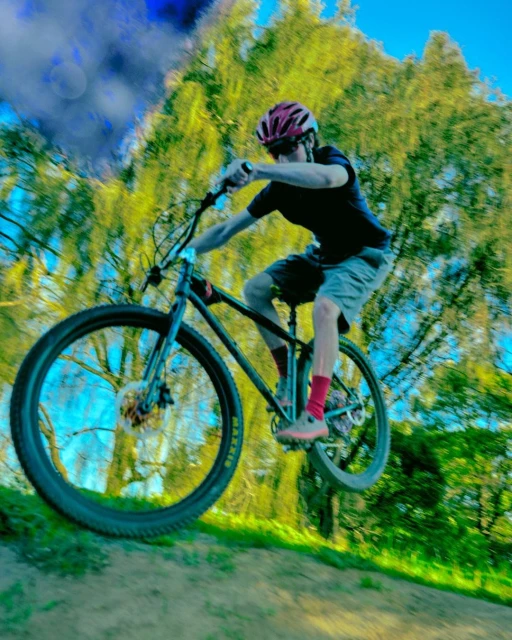 A cyclist wearing a helmet is in mid-air while riding a mountain bike, trees are visible in the background on a sunny day.