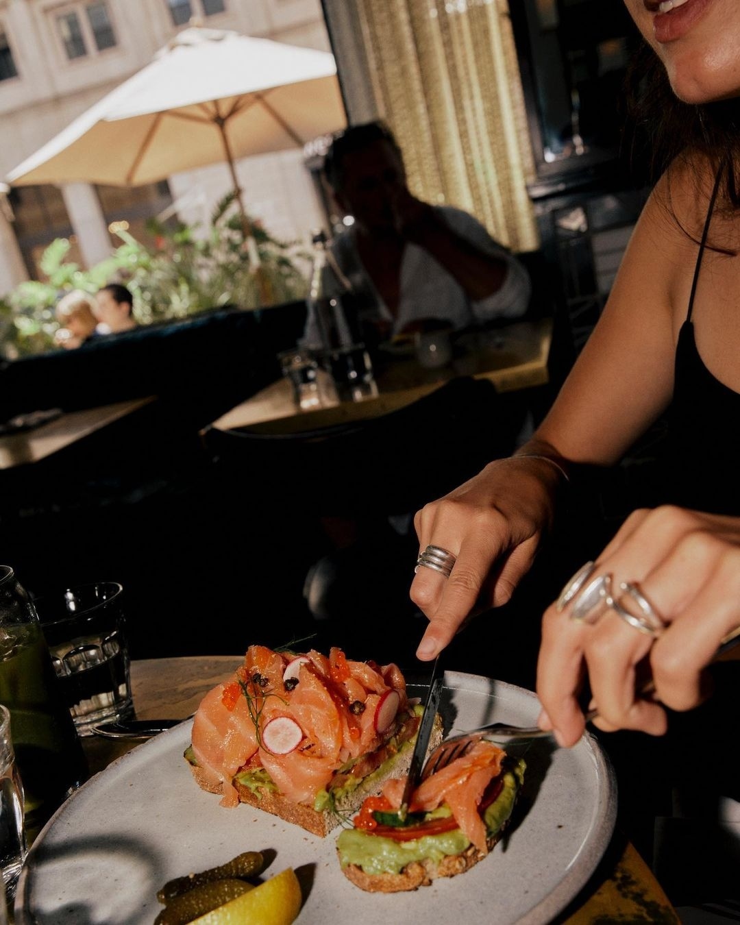 Woman cuts open-faced salmon and avocado sandwiches topped with red caviar at indoor restaurant, served with lemon and pickles.