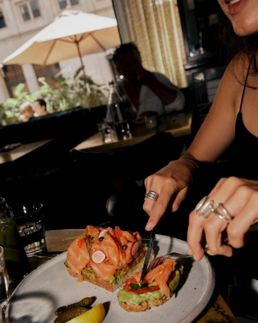 Woman cuts open-faced salmon and avocado sandwiches topped with red caviar at indoor restaurant, served with lemon and pickles.