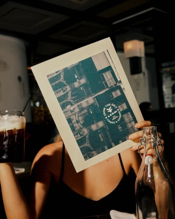 Person at a cafe holding a menu in front of their face, drink in hand, and a glass bottle on the table, with overhead aerial photography.