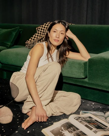 Woman seated on the floor leaning against a green velvet sofa, wearing a white tank top and beige trousers, photographed by Charlie Hawks.