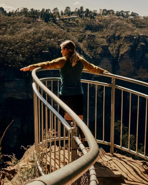 Woman on scenic overlook with outstretched arms, enjoying a view of Blue Mountains National Park in Australia on a sunny day.
