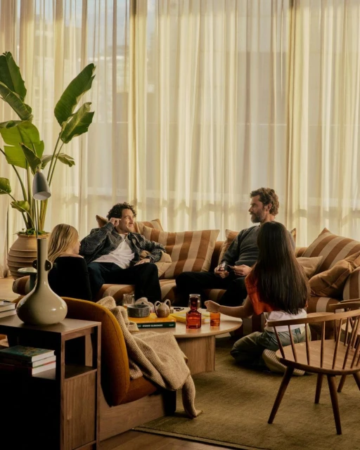 Four people relaxing in living room with large window, sofa, chairs, plants, and coffee table holding pitcher, glasses, books, and ceramics.