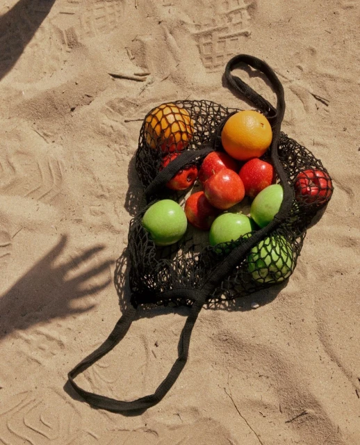 Black string bag filled with oranges, red apples, and green apples sitting on the sand, with a person's hand casting a shadow.