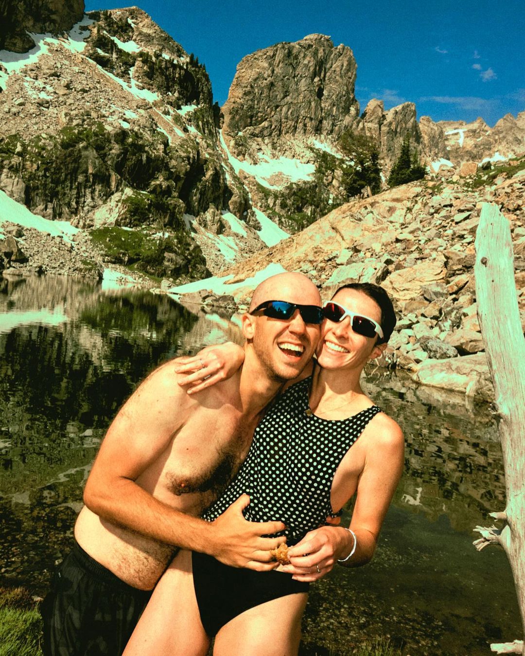 Couple embraces near mountain lake under blue sky. Adult male is shirtless with sunglasses, adult female wears polka-dot swimsuit.