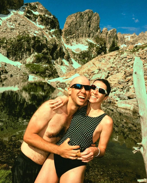 Couple embraces near mountain lake under blue sky. Adult male is shirtless with sunglasses, adult female wears polka-dot swimsuit.