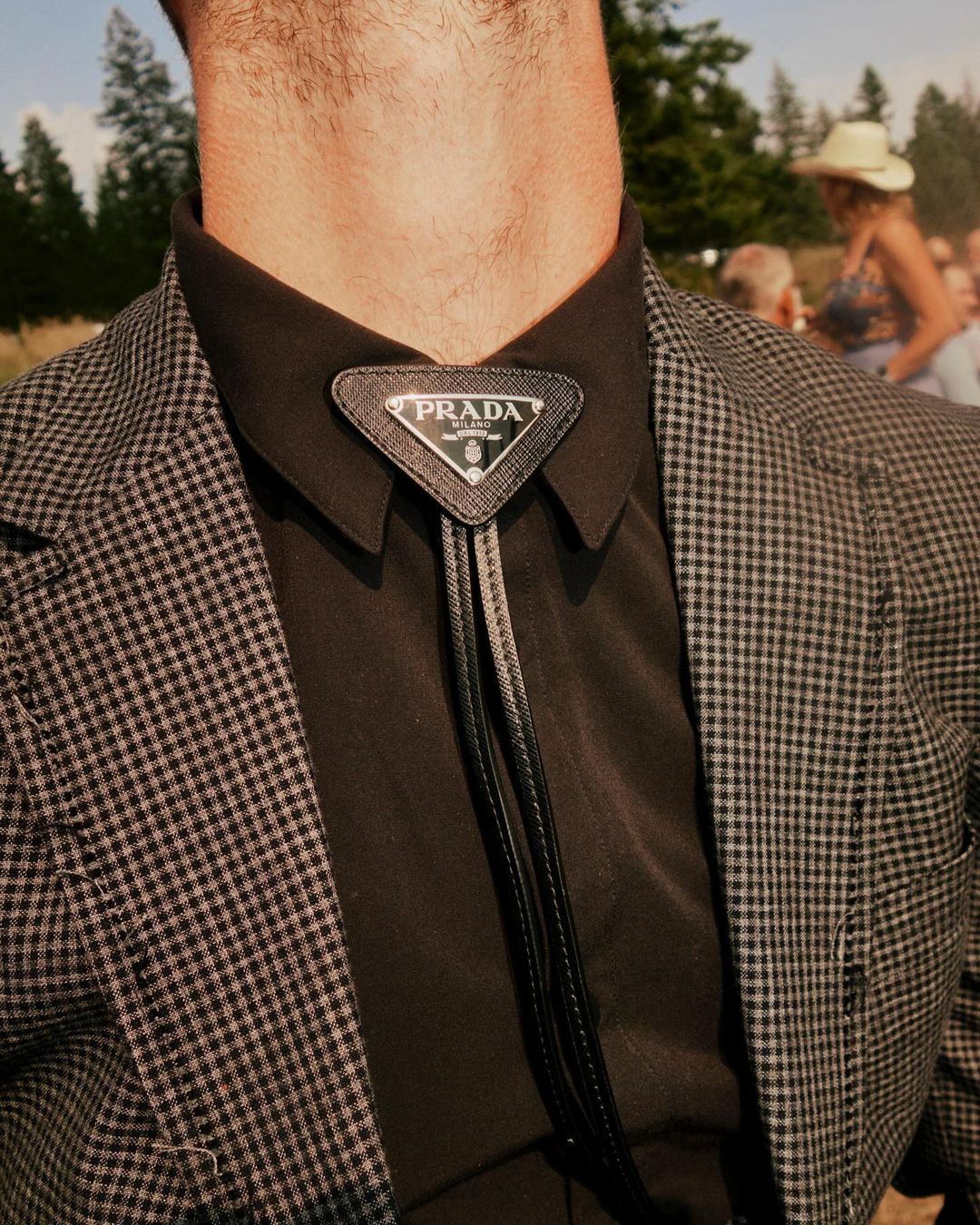 Man wearing a Prada bolo tie, black dress shirt, and grey checkered suit jacket, standing in a field with blurred figures in background.