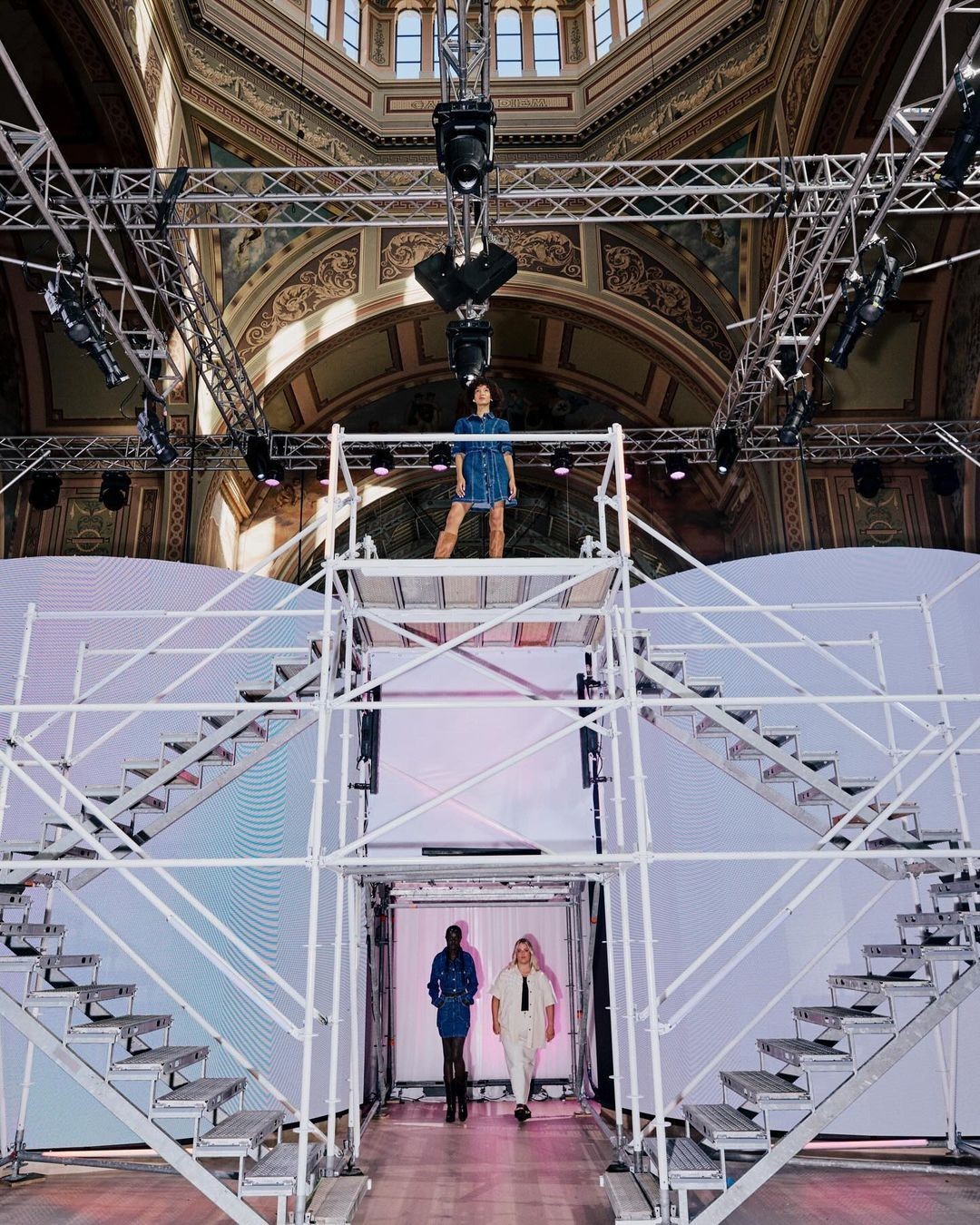 Fashion show catwalk featuring a female model on a scaffolding platform and two models walking on the runway in an ornate interior.