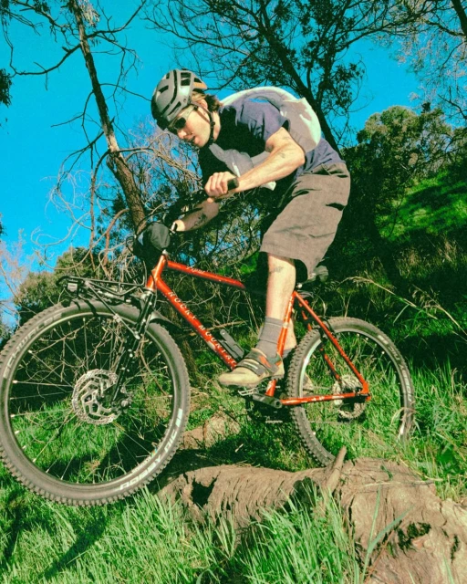Adult male mountain biker wearing helmet and sunglasses riding red mountain bike through a grassy, tree-lined trail under blue sky.