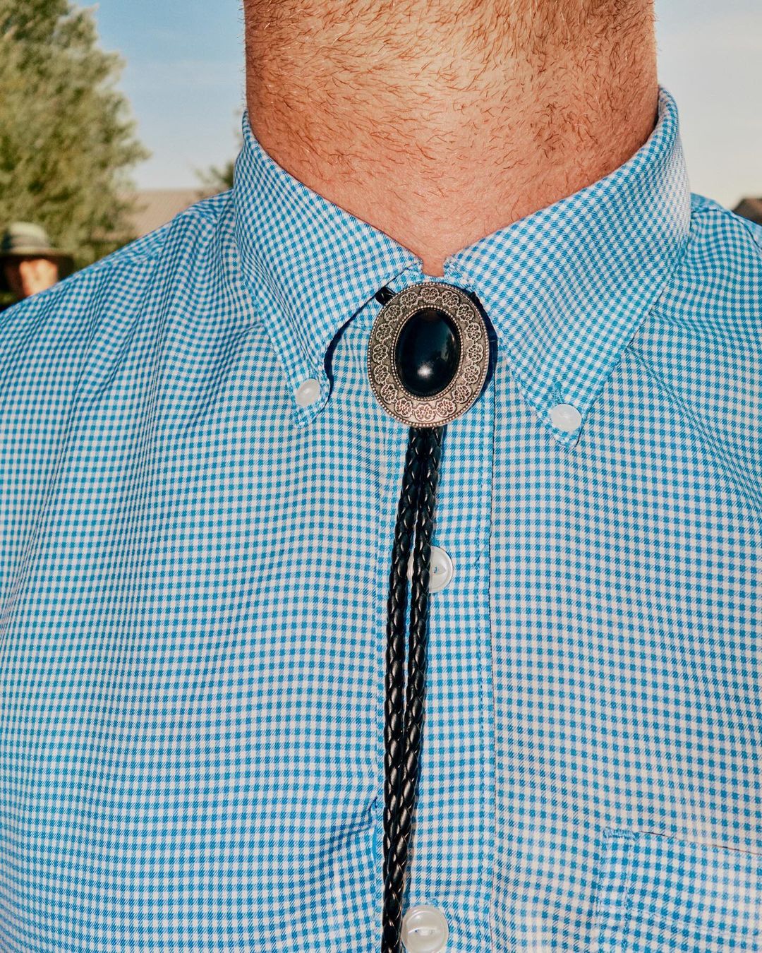 Close-up of a man wearing a light blue checked shirt and a bolo tie with a silver oval and black stone outdoors on a sunny day.