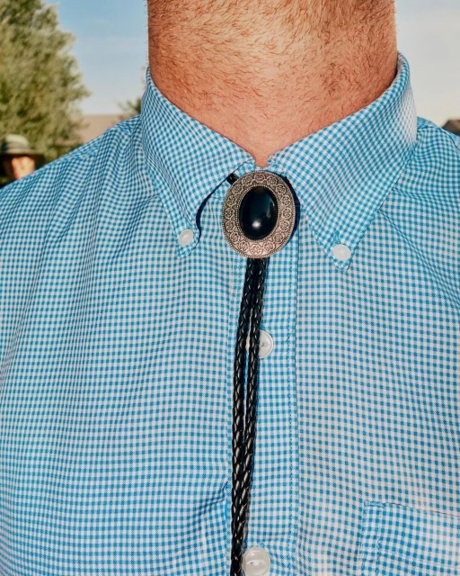 Close-up of a man wearing a light blue checked shirt and a bolo tie with a silver oval and black stone outdoors on a sunny day.