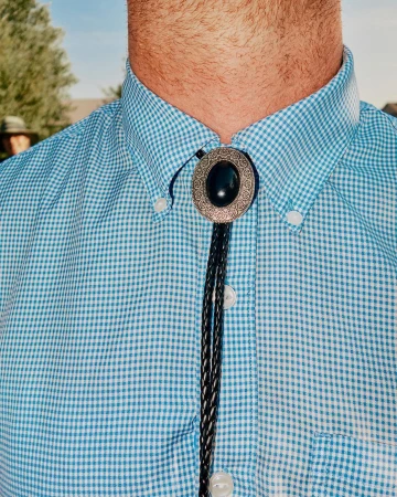 Close-up of a man wearing a light blue checked shirt and a bolo tie with a silver oval and black stone outdoors on a sunny day.