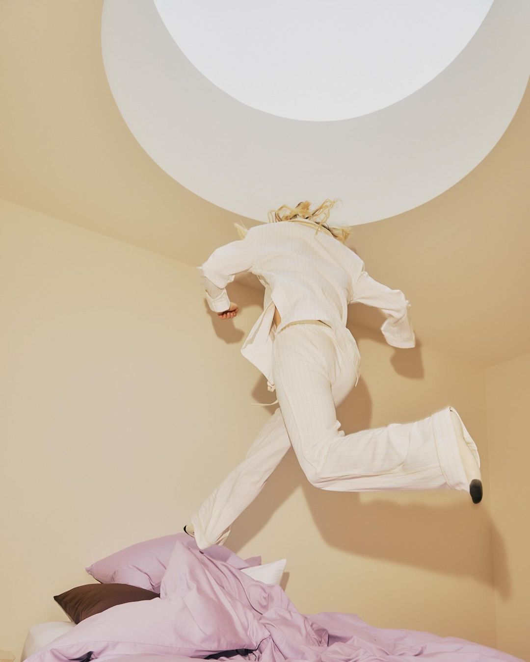 Person jumping over bed in all white in a room with a circular light in the ceiling. The scene is captured from a low angle.