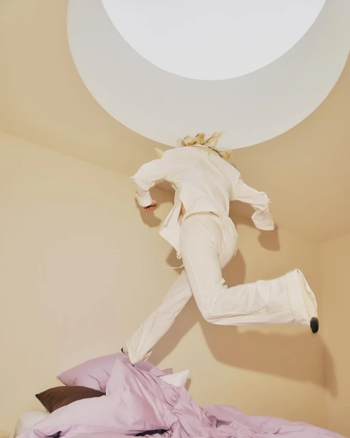 Person jumping over bed in all white in a room with a circular light in the ceiling. The scene is captured from a low angle.