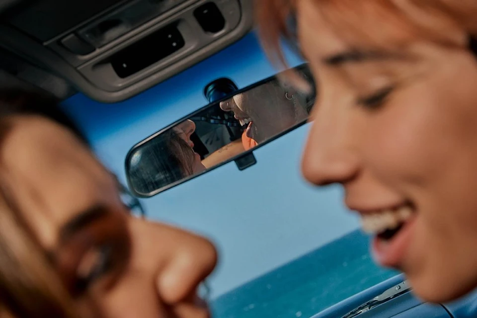 Close-up shot inside a car features two females in blurry foreground. They're reflected in the rearview mirror against blue sky and ocean.