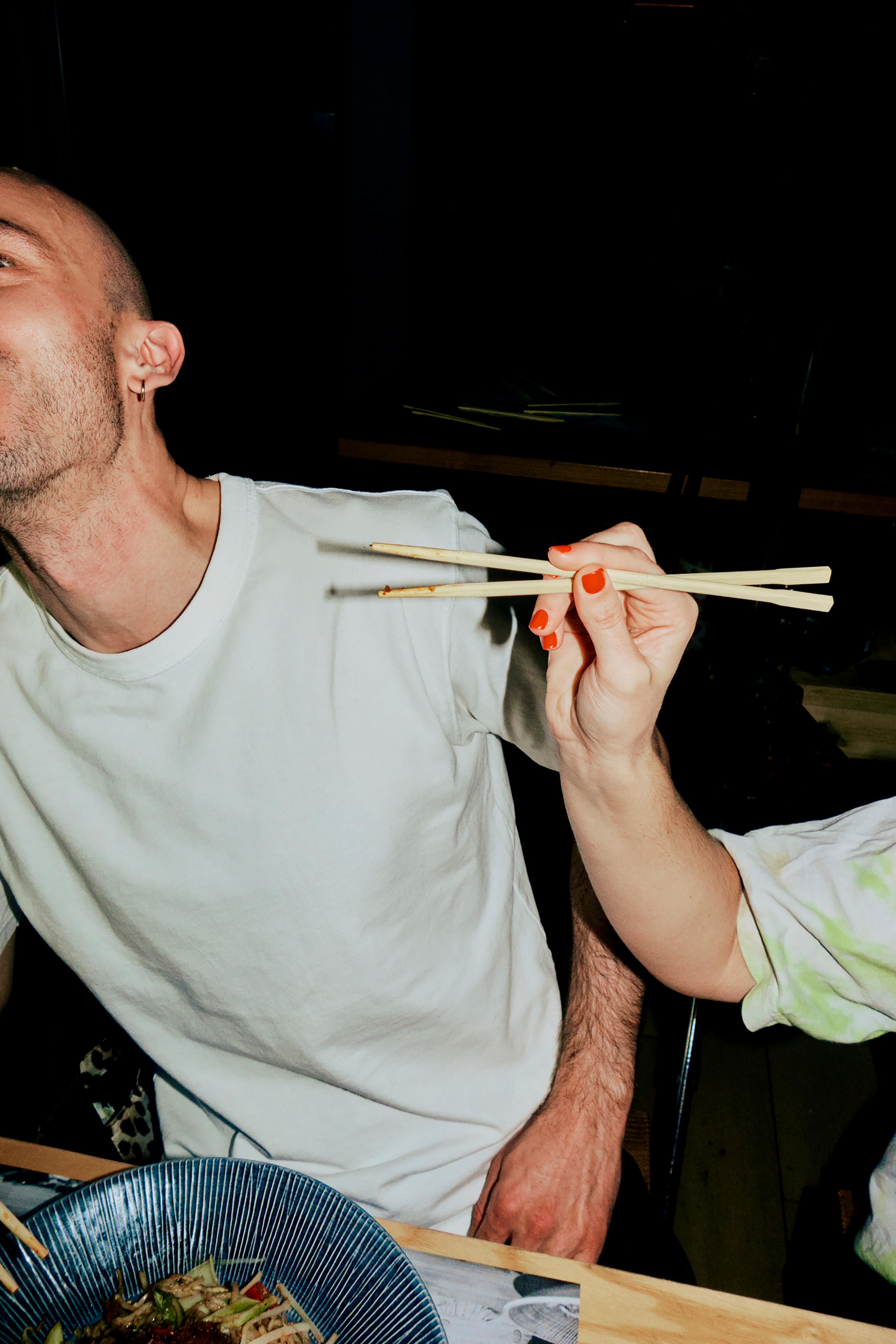 A bald man eats Asian food with chopsticks, held by another person with red nail polish, in an indoor restaurant setting.