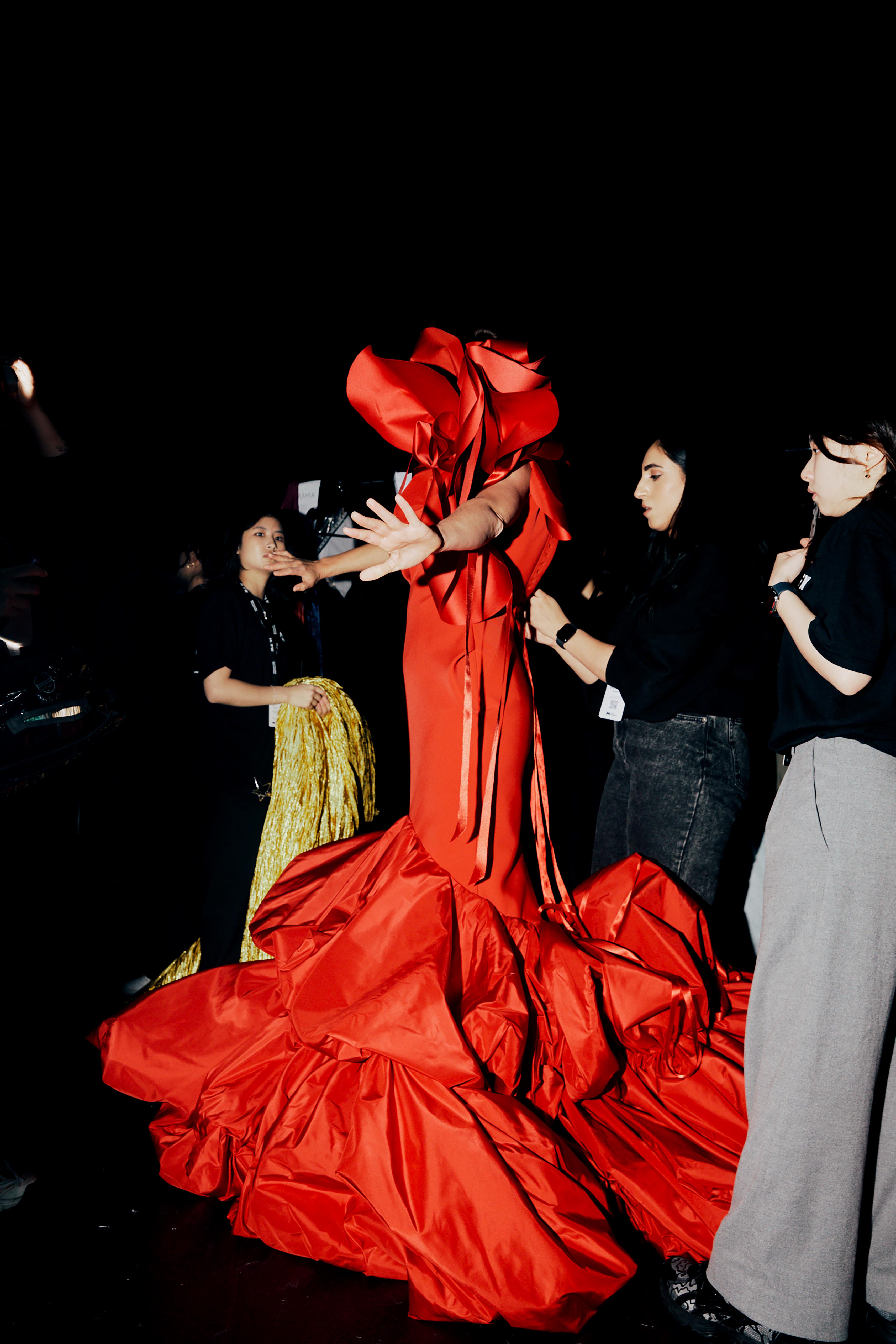 Runway fashion shot featuring a model in an elaborate, voluminous red gown surrounded by a small group of fashion support staff.
