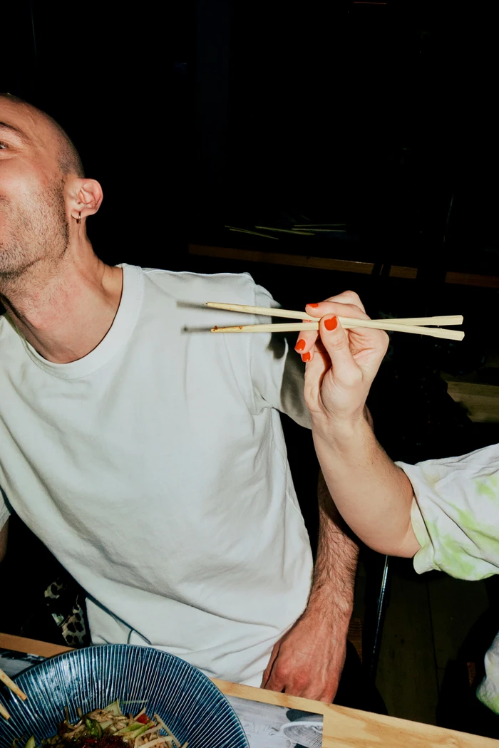 Close-up portrait of a man in a white t-shirt holding chopsticks over a blue bowl of noodles in a restaurant setting.