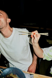 Close-up portrait of a man in a white t-shirt holding chopsticks over a blue bowl of noodles in a restaurant setting.