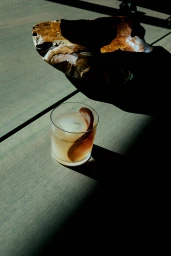 A rocks glass with an alcoholic drink and ice sits on a wooden table alongside a carved wooden object under dramatic hard light.