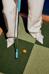 Person in white trousers and pointed shoes holding a blue putter next to an orange golf ball on a green turf carpet