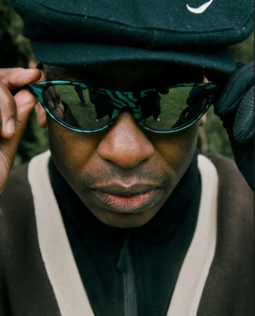Close-up portrait of a Black man adjusting teal patterned sunglasses, wearing a dark cap and brown collared zip jacket.
