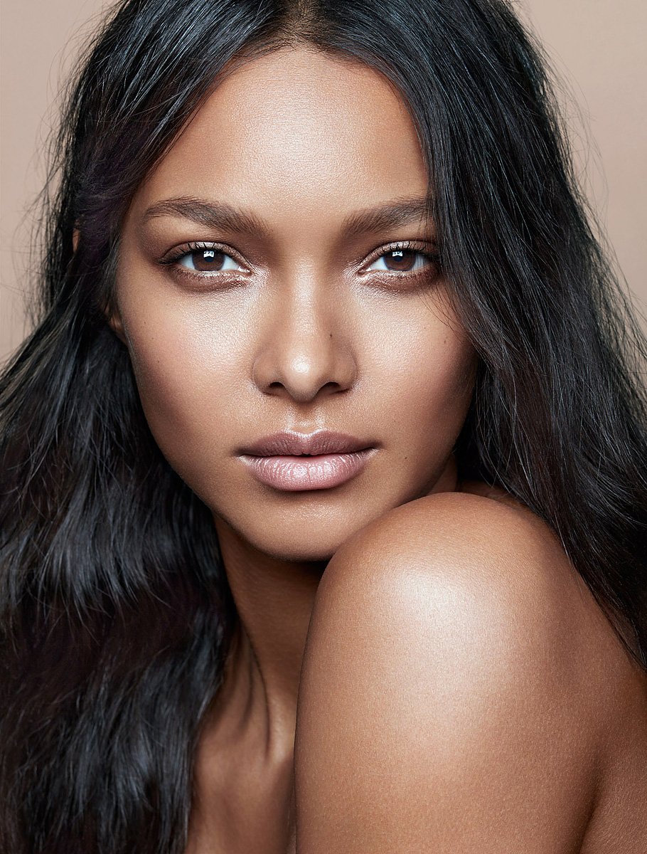 Close-up portrait of a beautiful adult female model with dark hair and brown eyes, posing in a beauty shot against a tan backdrop.
