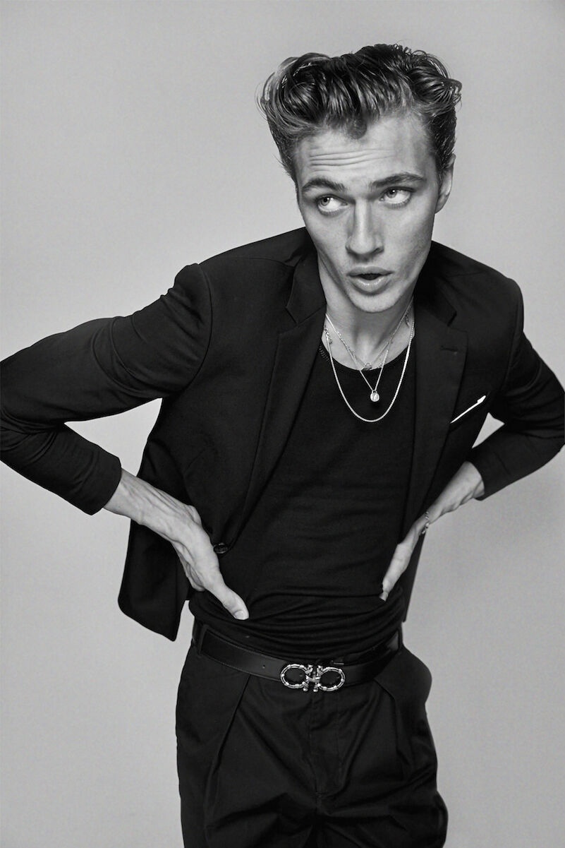 Monochrome portrait of a young adult man in a black suit, black shirt, and silver necklaces, posing with hands on his belt.