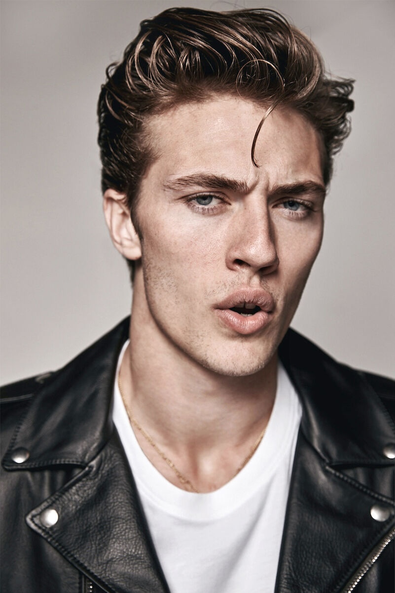 Portrait of a young adult man with light brown wavy hair, wearing a black leather jacket and white t-shirt, against a grey background.