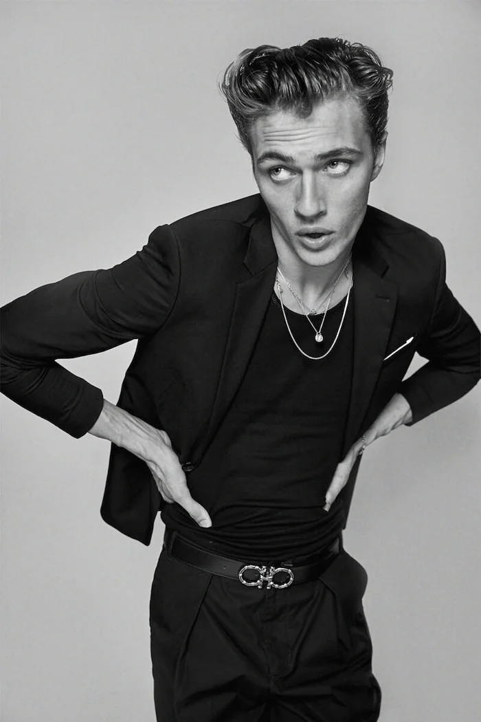 Monochrome fashion portrait of a young man wearing a black blazer, t-shirt, and leather belt against a neutral background.
