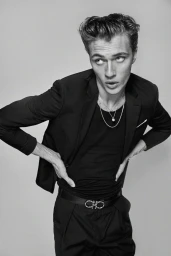 Monochrome fashion portrait of a young man wearing a black blazer, t-shirt, and leather belt against a neutral background.