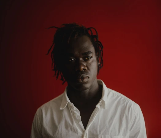Portrait of a Black man with dreadlocks wearing a white button-down shirt against a dark red background by photographer Yeoseop Yoon
