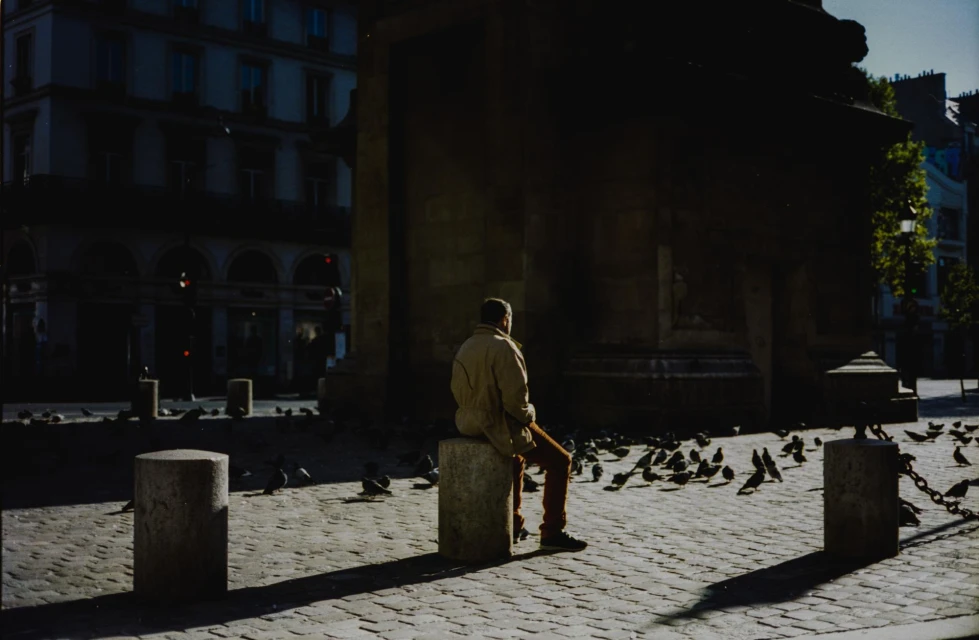 Man in coat sits on stone bollard surrounded by pigeons on a brick plaza in the city during daytime; buildings in the background.